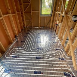 BUDUJE-GRUPA Spółka z ograniczoną odpowiedzialnością - Instalacja ogrzewania podłogowego w domu szkieletowym, rury ułożone na folii z siatką, widoczne drewniane elementy konstrukcji dachu i okno dachowe.