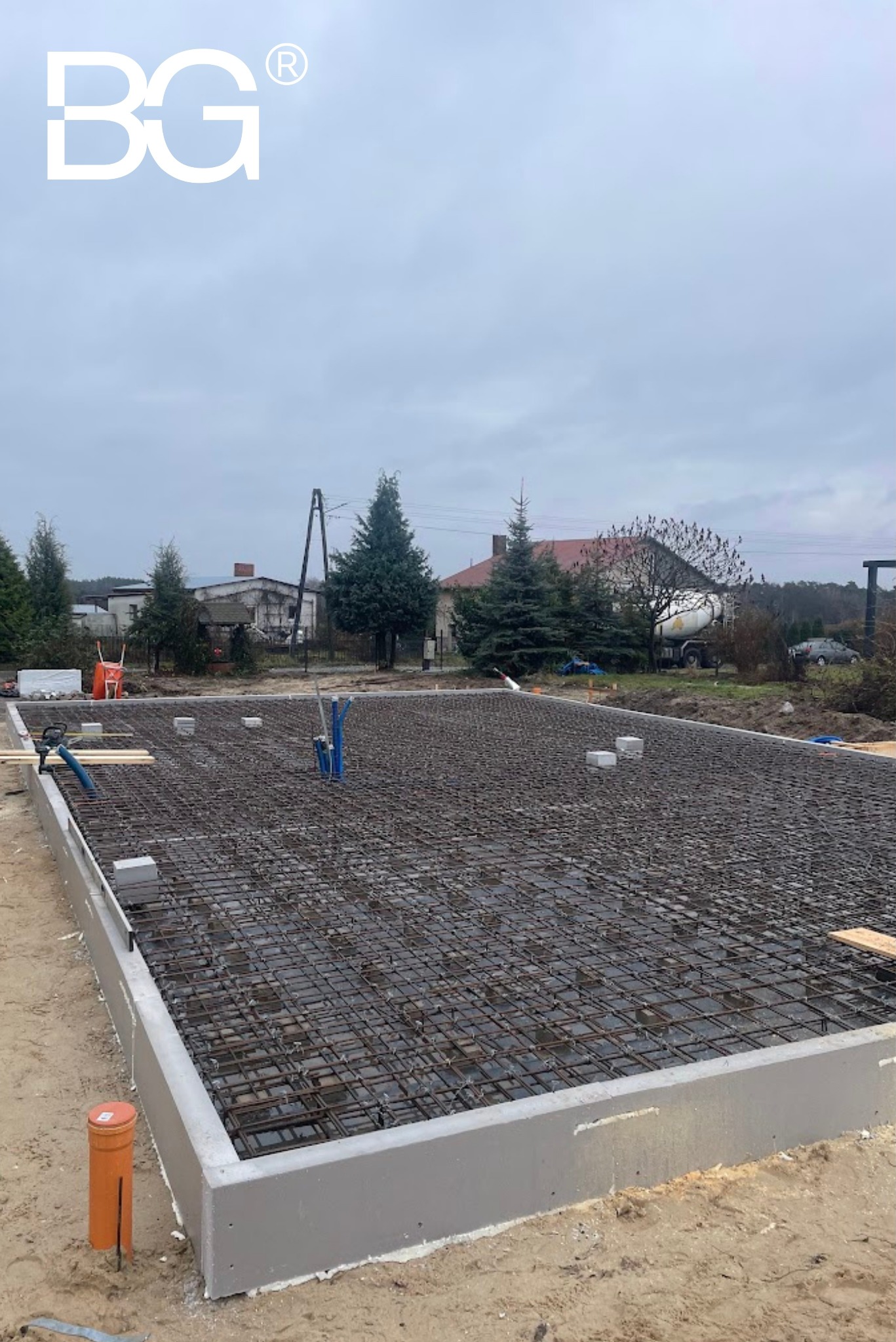 Zbrojenie fundamentów pod budowę domu, widoczne szalunki, wystające rury instalacyjne. W tle domy i betoniarka. Pochmurny dzień.