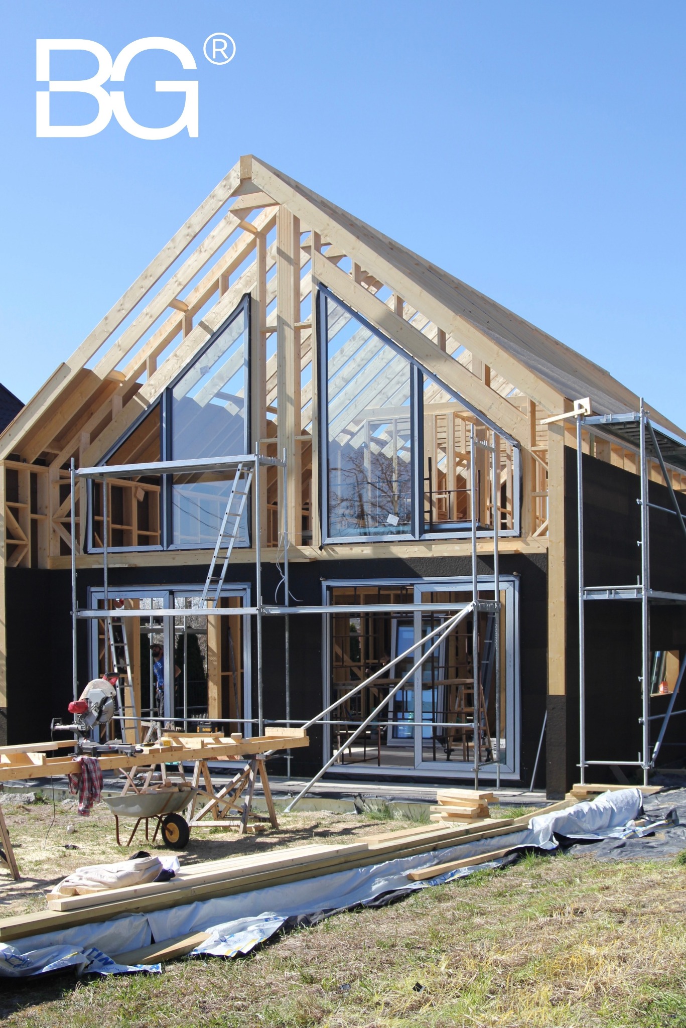 Wznoszenie domu z drewnianą konstrukcją szkieletową, widoczne rusztowania i duże okna. Prace w toku, elementy konstrukcyjne na pierwszym planie.