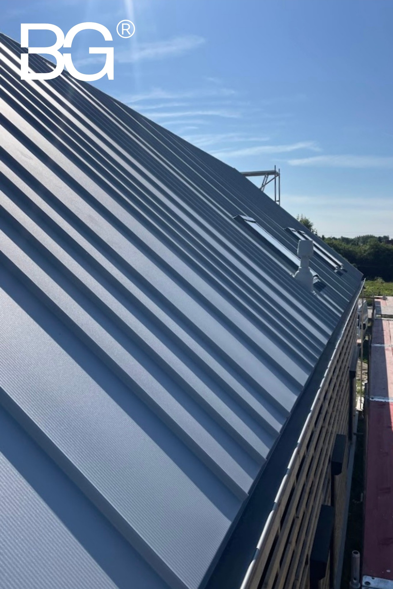 Szary dach z blachy trapezowej z oknami dachowymi i kominkiem wentylacyjnym, widok z dołu pod kątem, błękitne niebo w tle, logo firmy w lewym górnym rogu.