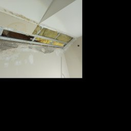 Rafał Juszczak - Odsłonięty fragment sufitu z widoczną konstrukcją metalową, izolacją i śladami wilgoci na ścianie. Widoczne uszkodzenia i elementy wymagające naprawy.