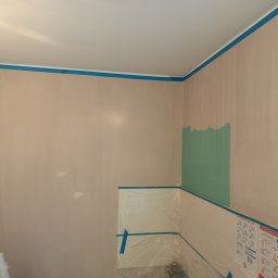 Rafał Juszczak - Przygotowanie do malowania: ściana z fragmentem pomalowanym na zielono, zabezpieczona folią i taśmą malarską, widoczne narzędzia malarskie.