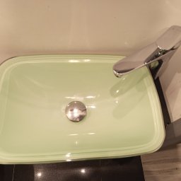 Rafał Juszczak - Jasnozielona, prostokątna umywalka z chromowaną baterią i odpływem, zamontowana na czarnym blacie. Widok z góry, oświetlenie ciepłe.