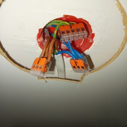 Rafał Juszczak - Połączenie przewodów elektrycznych (brązowych, niebieskich, zielono-żółtych) za pomocą pomarańczowych złączek WAGO 221, wystających z czerwonej puszki instalacyjnej w okrągłym otworze w białym...