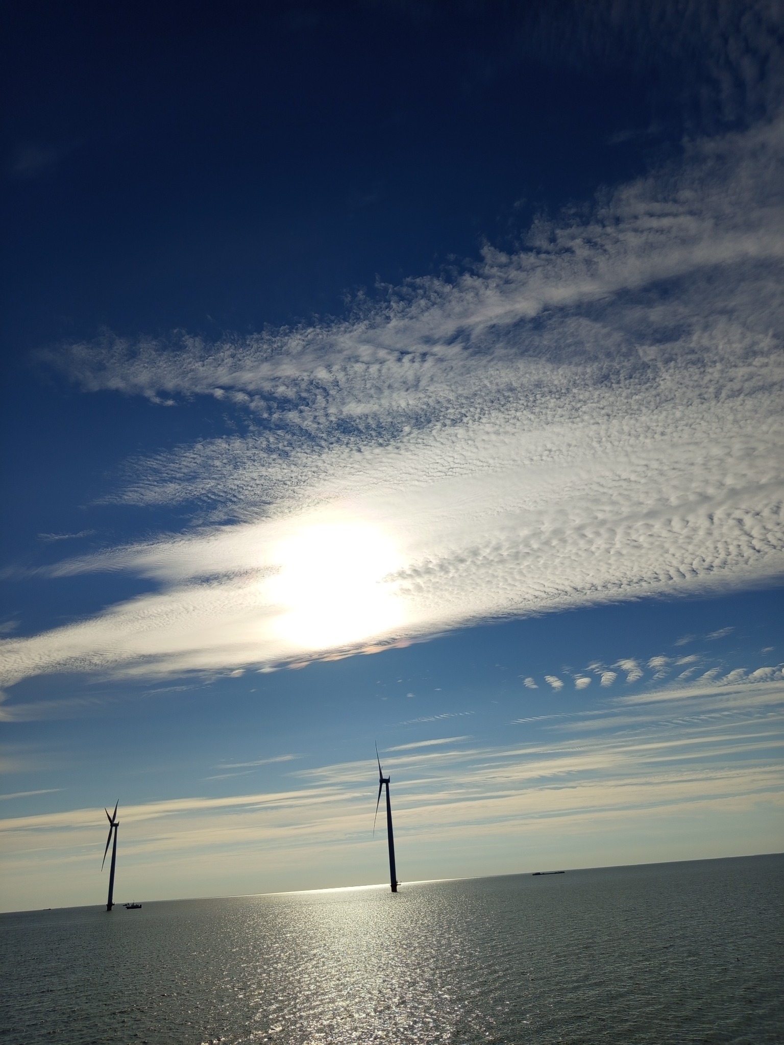 Dwie turbiny wiatrowe na morzu pod błękitnym niebem z chmurami, odbijające słońce w wodzie. Perspektywa z dołu, podkreślająca wysokość konstrukcji.