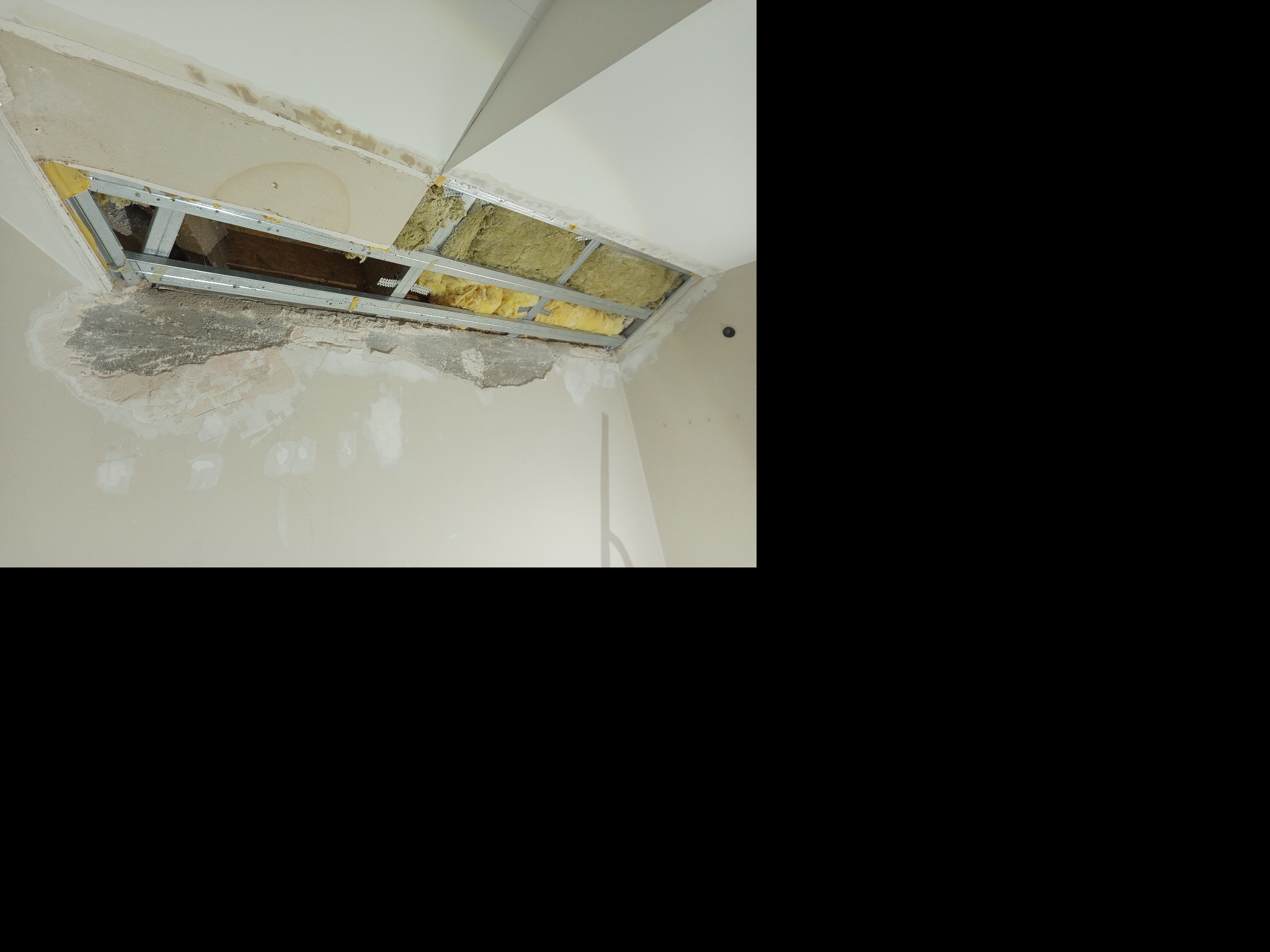 Odsłonięty fragment sufitu z widoczną konstrukcją metalową, izolacją i śladami wilgoci na ścianie. Widoczne uszkodzenia i elementy wymagające naprawy.