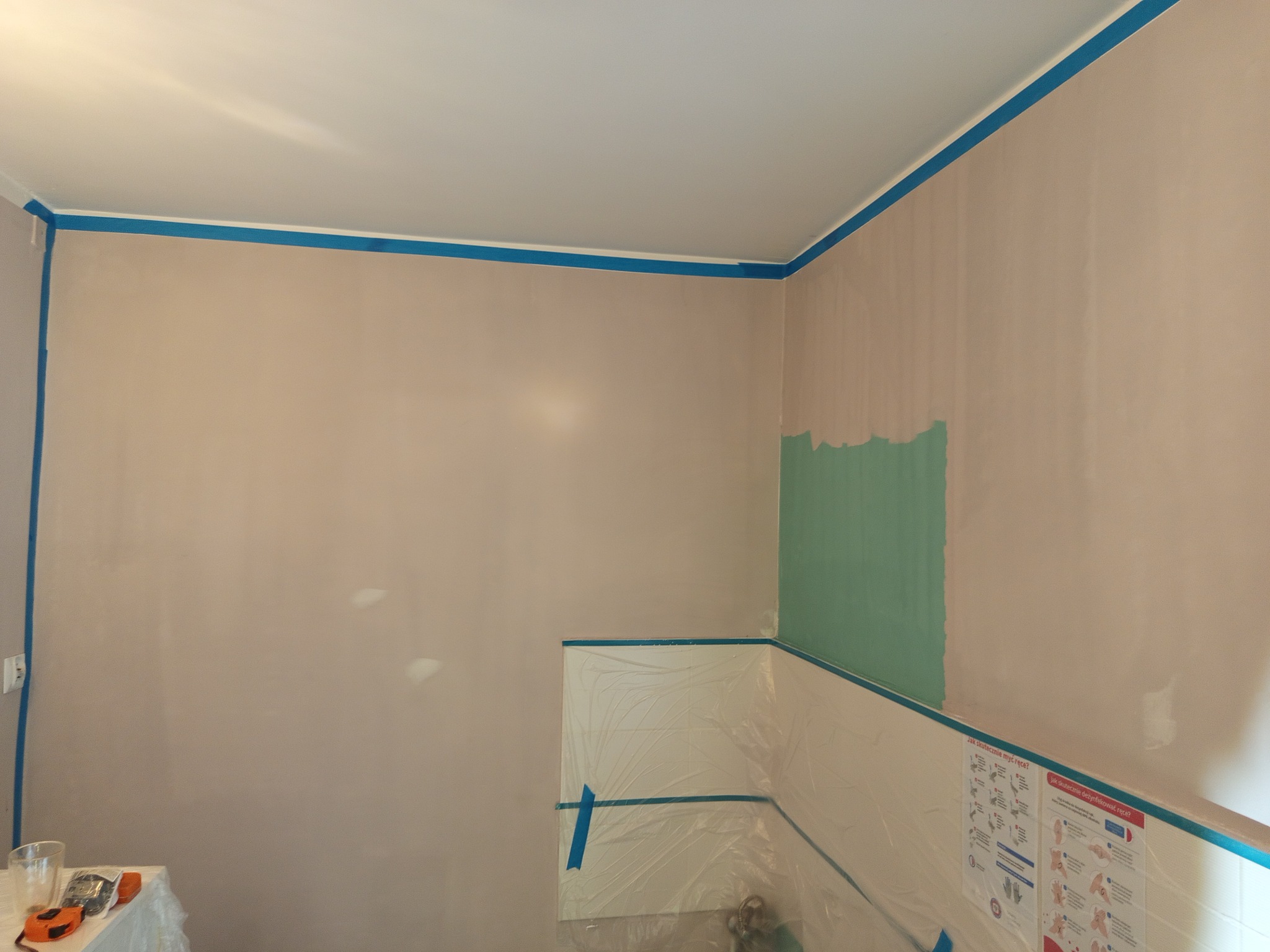 Przygotowanie do malowania: ściana z fragmentem pomalowanym na zielono, zabezpieczona folią i taśmą malarską, widoczne narzędzia malarskie.