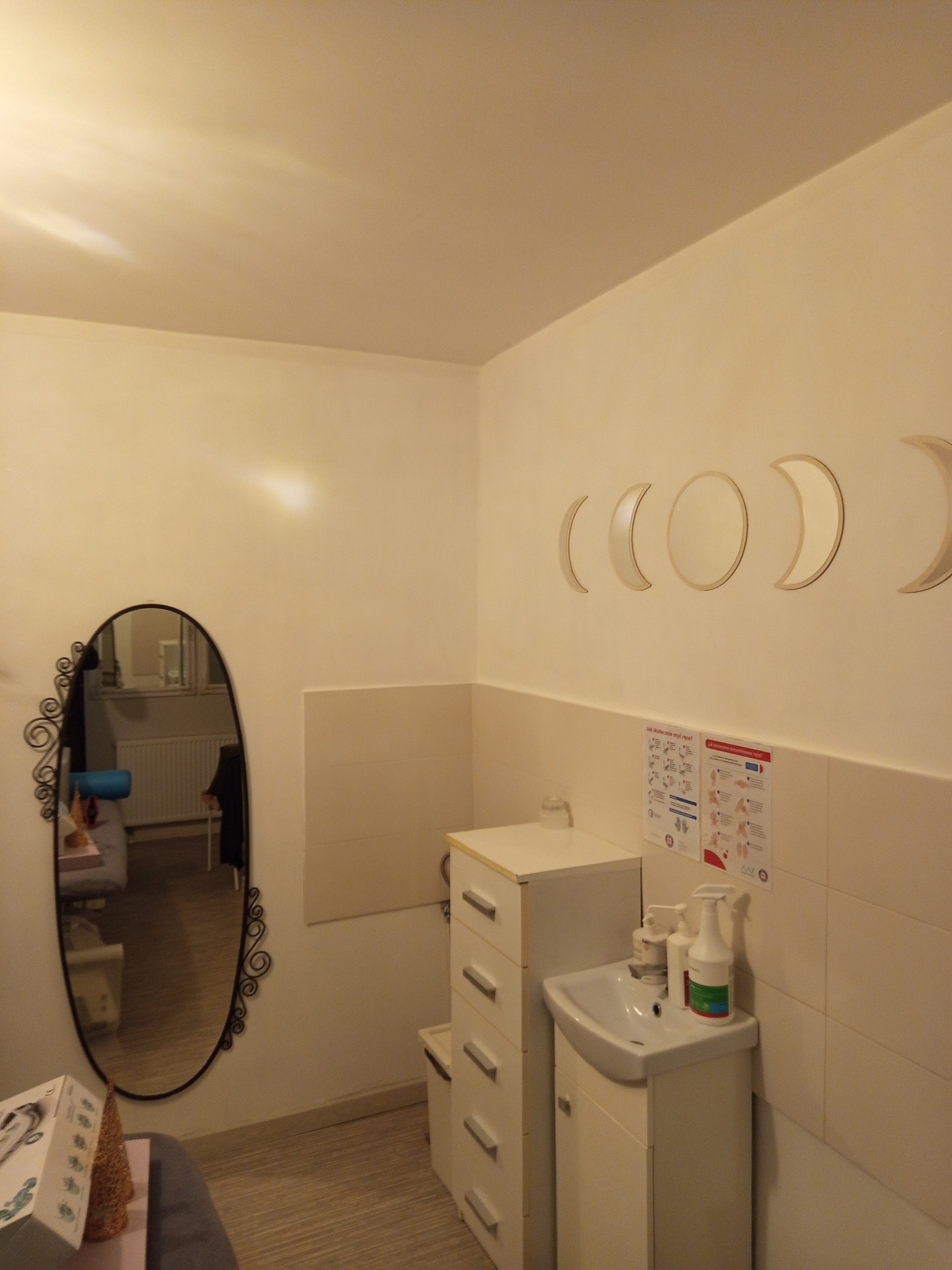 Jasne wnętrze gabinetu kosmetycznego z umywalką, szafką i dekoracyjnym lustrem. Na ścianie instrukcja mycia rąk i lustrzane fazy księżyca.