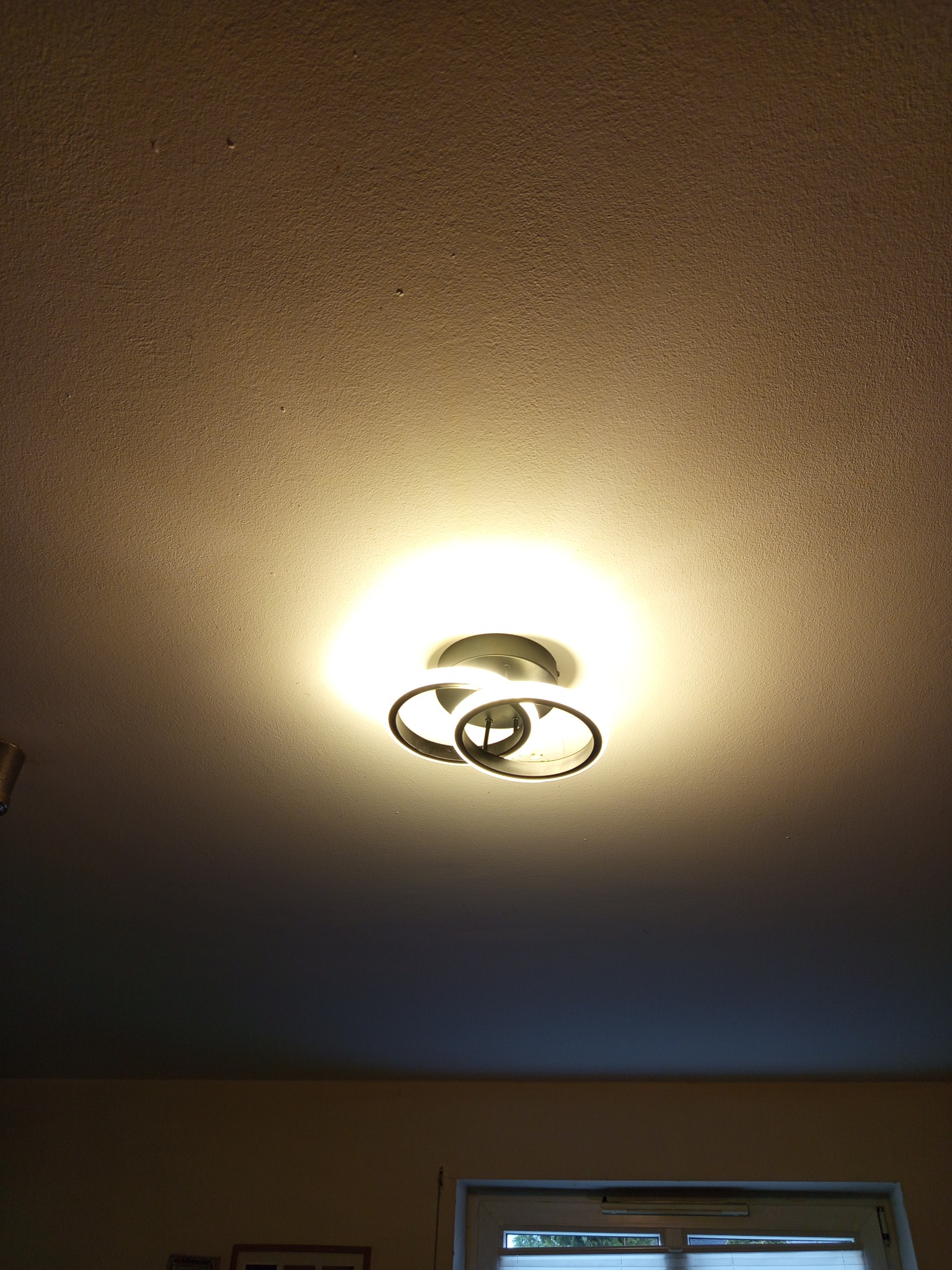 Nowoczesna lampa sufitowa z trzema metalowymi okręgami, emitująca ciepłe światło na teksturowany sufit. Widok z dołu, fragment ściany i okna w tle.