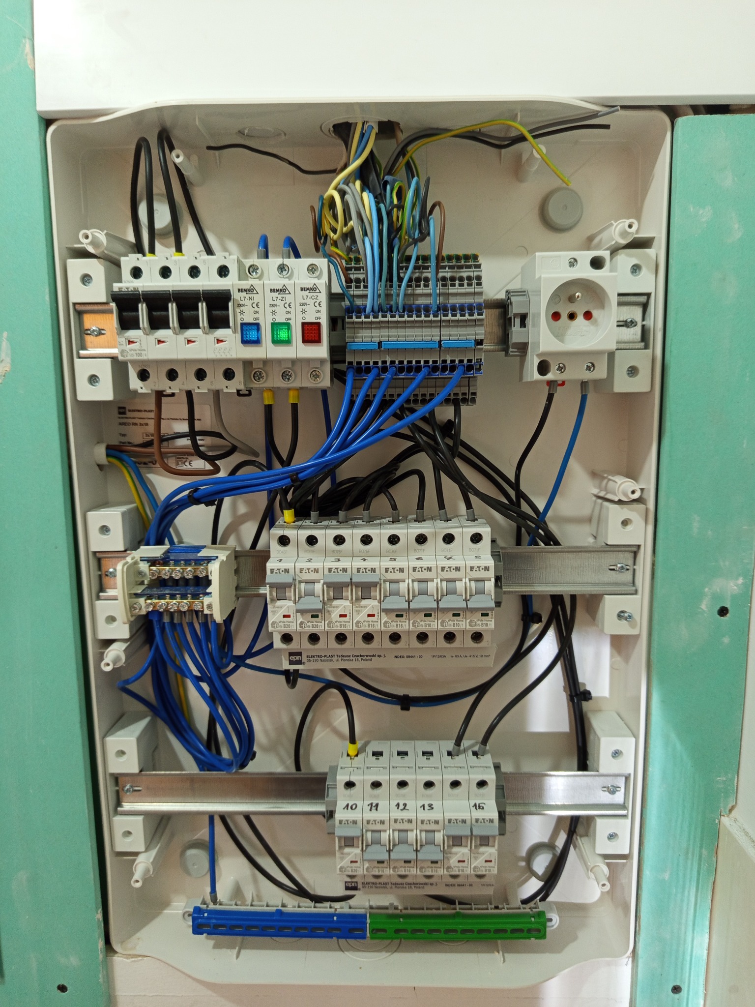 Rozdzielnica elektryczna po montażu, widoczne bezpieczniki, przewody i gniazdo. Instalacja w białej obudowie, zamontowana na ścianie z płyt gipsowo-kartonowych.
