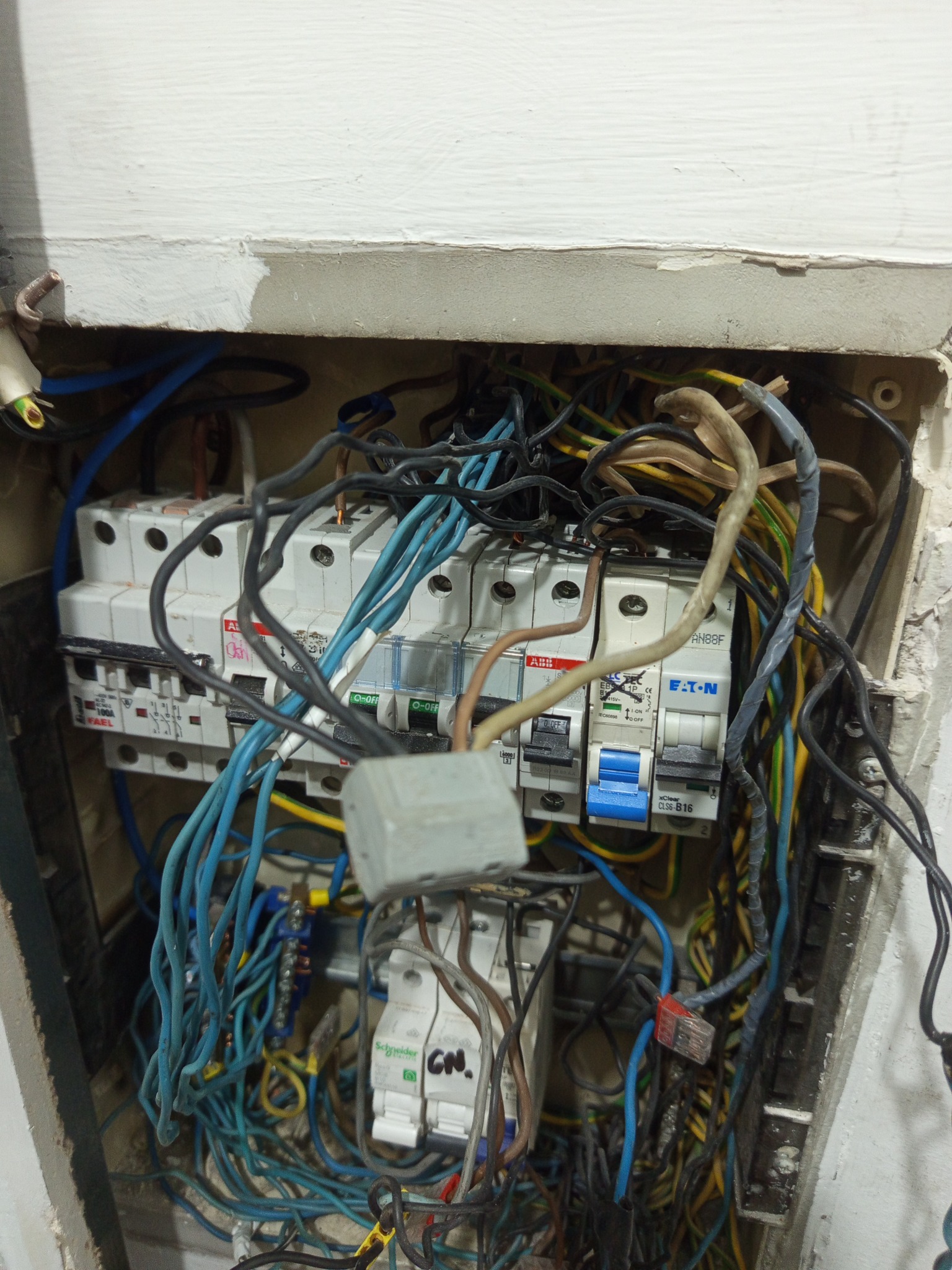 Otwarta skrzynka elektryczna z widocznymi bezpiecznikami i plątaniną kabli w różnych kolorach, umieszczona w ścianie. Widoczne ślady po szpachlowaniu.