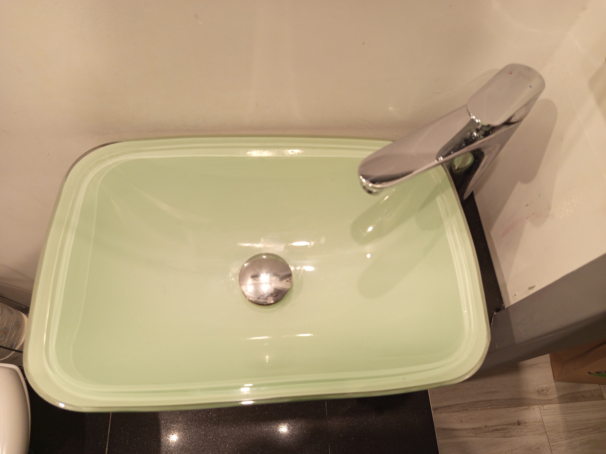 Jasnozielona, prostokątna umywalka z chromowaną baterią i odpływem, zamontowana na czarnym blacie. Widok z góry, oświetlenie ciepłe.