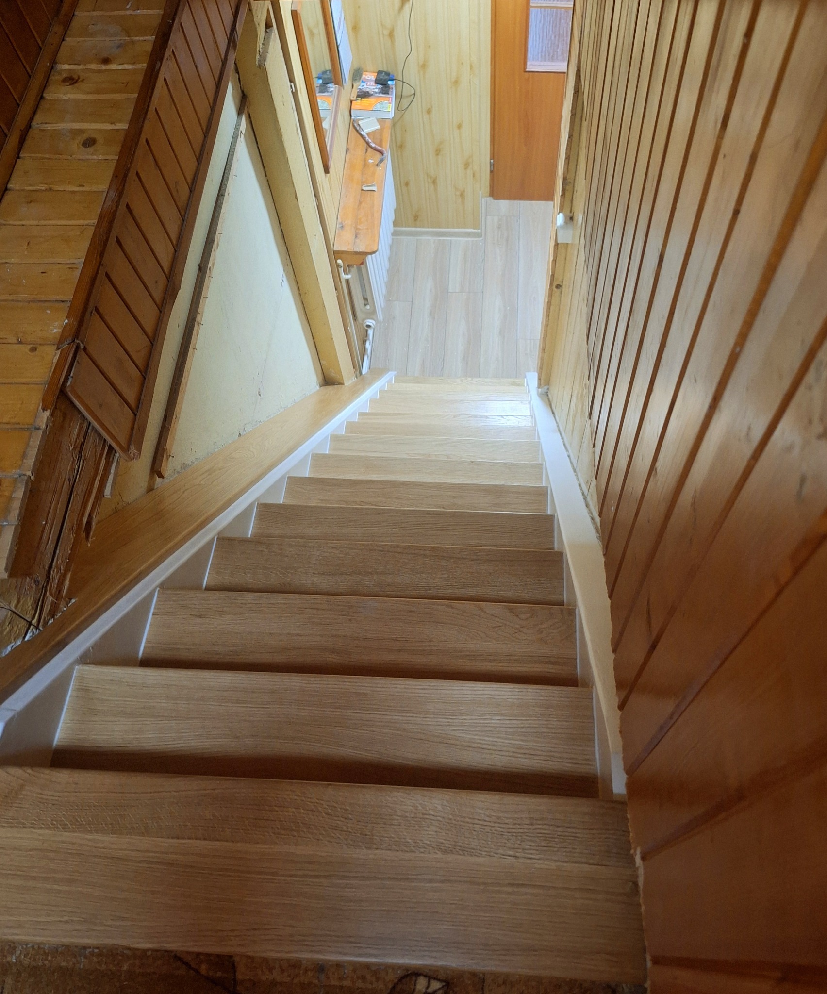 Jasne, drewniane schody z białymi podstopnicami, widziane z góry. Ściany wykończone boazerią. Widoczny fragment podłogi i drzwi na górze schodów.