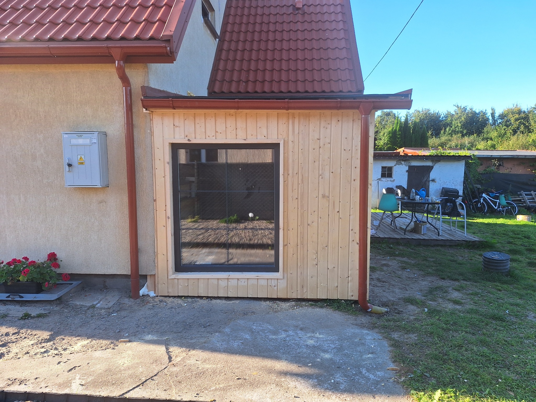Nowa, drewniana dobudówka z oknem, przylegająca do budynku z czerwoną dachówką. Widoczne rynny i skrzynka elektryczna na ścianie. Tło: zieleń i zabudowania gospodarcze.