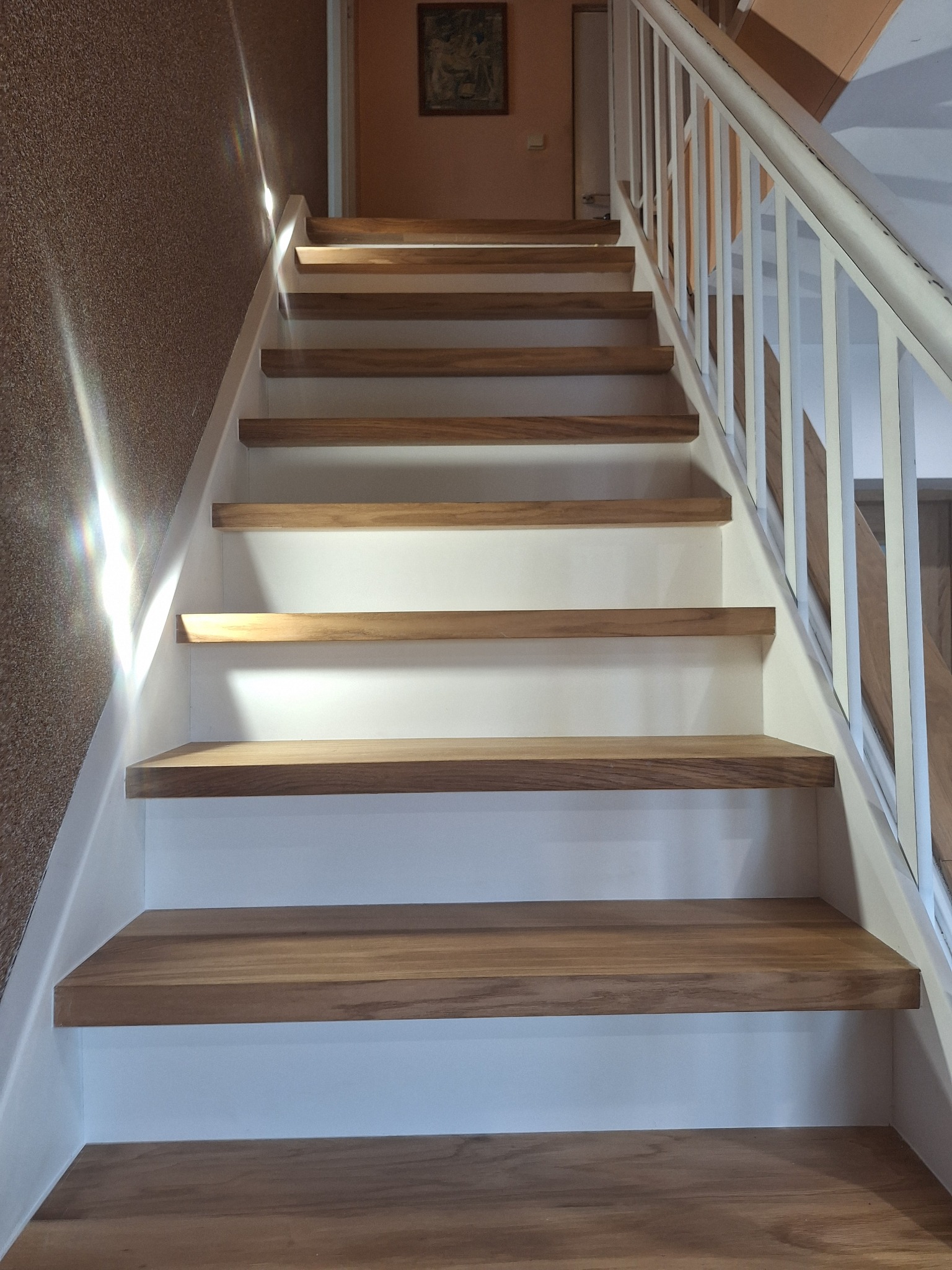 Drewniane schody z białymi podstopnicami i białą balustradą, oświetlone naturalnym światłem. Widok z dołu na górę, z widoczną fakturą ściany po lewej stronie.