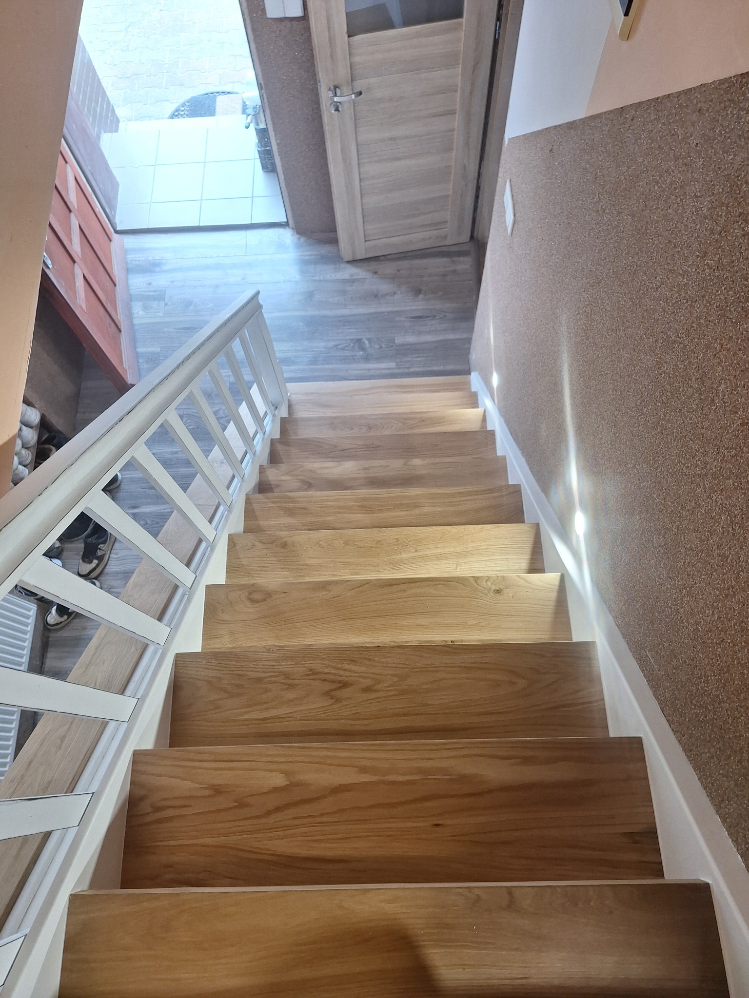 Drewniane schody z białymi barierkami, widok z góry. Naturalne oświetlenie podkreśla strukturę drewna. Widoczny fragment drzwi i podłogi.