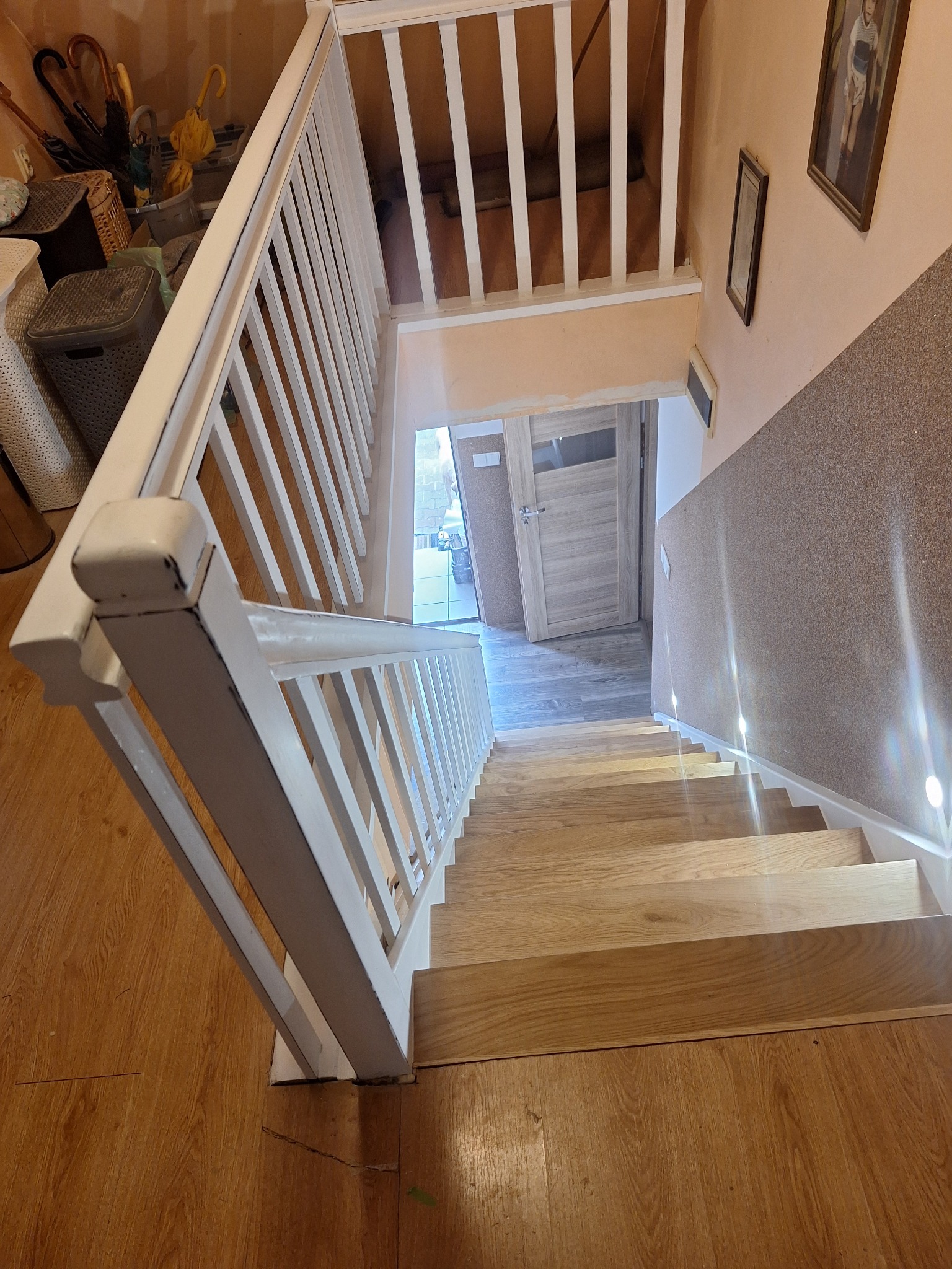 Jasne, drewniane schody z białymi balustradami, widziane z góry. Na ścianie wiszą obrazy, a w tle widać drzwi. Podłoga wykończona panelami.