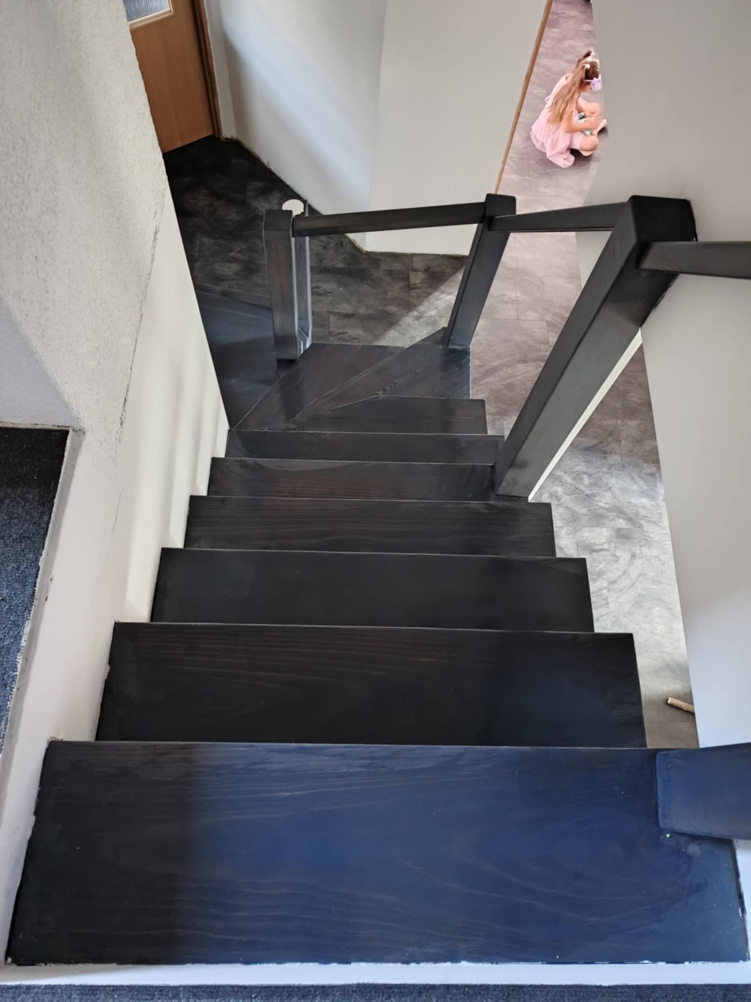 Ciemne, drewniane schody z metalową balustradą, widok z góry. Na górze schodów widoczna osoba siedząca na podłodze. Minimalistyczny design.