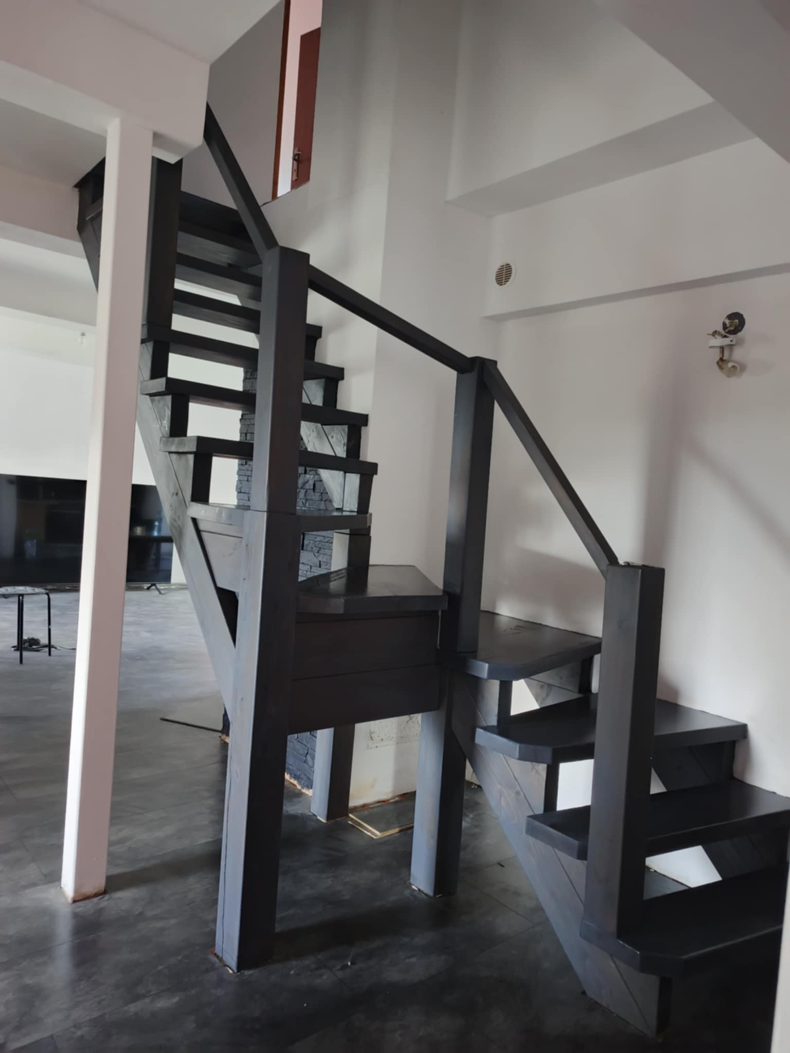 Ciemne, drewniane schody z balustradą w jasnym wnętrzu. Widoczne stopnie i elementy konstrukcyjne. Minimalistyczny design, nowoczesne wykończenie. Ujęcie z dołu, perspektywa podkreśla wysokość.