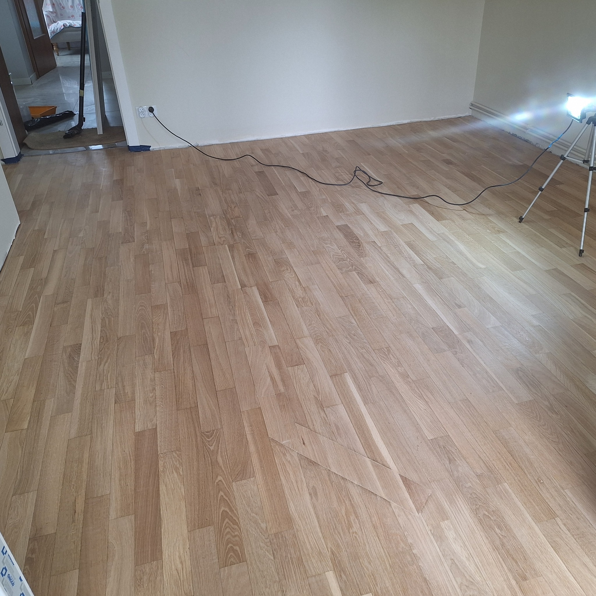 Jasne panele podłogowe ułożone w pokoju, widoczny kabel i lampa oświetleniowa. Świeżo położona podłoga w pustym pomieszczeniu.