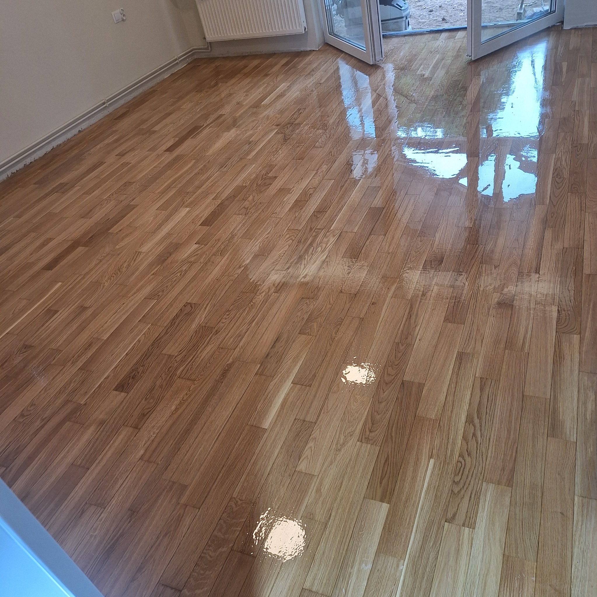 Błyszcząca, świeżo polakierowana podłoga parkietowa z wyraźnym rysunkiem drewna, odbijająca światło z otwartych drzwi balkonowych. Widoczne ślady po lakierowaniu.