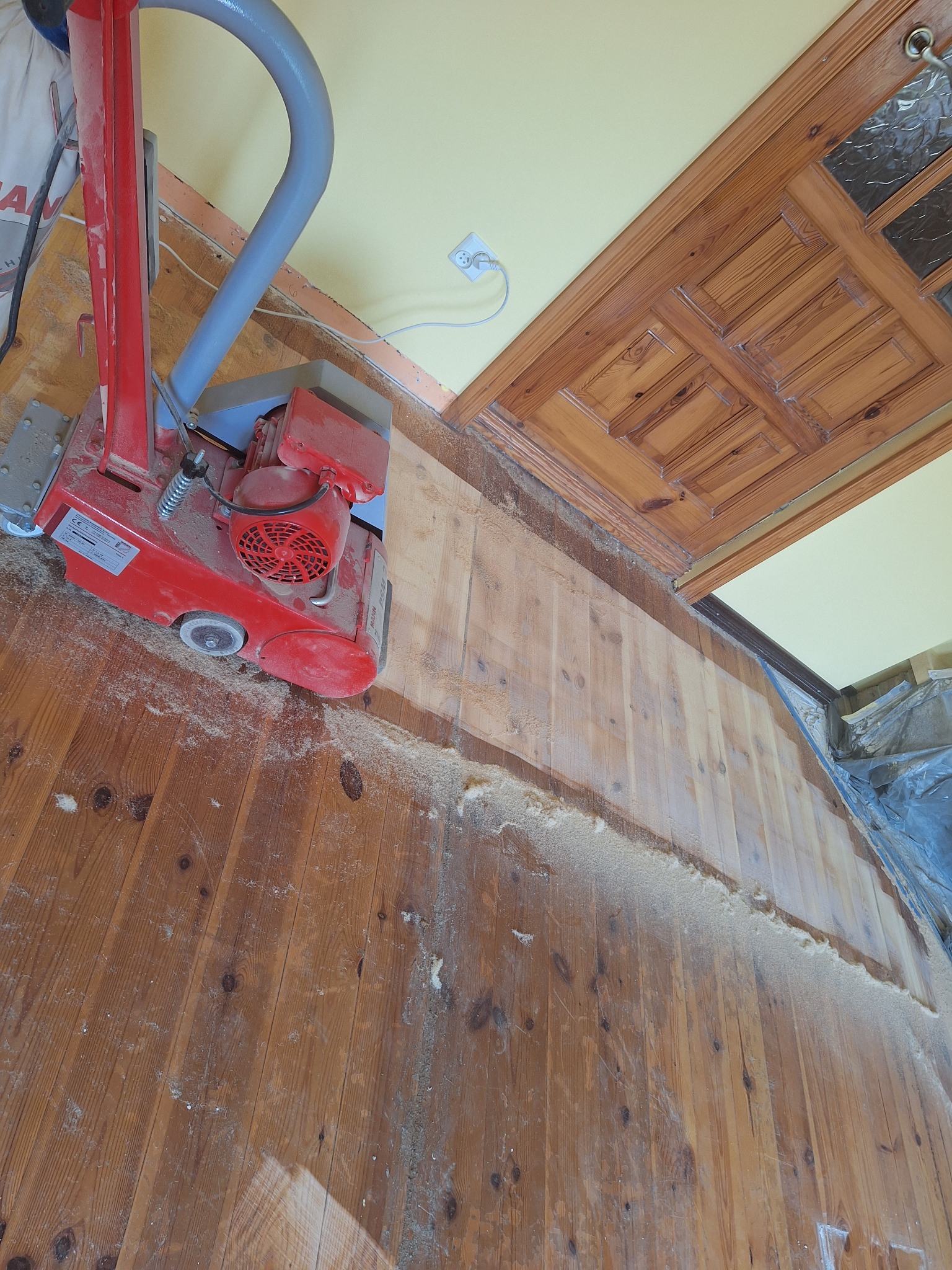 Czerwona cykliniarka w akcji na drewnianej podłodze w Pasłęku, widoczne ślady pyłu i obrobione deski. Ujęcie z góry, fragment drewnianego sufitu.