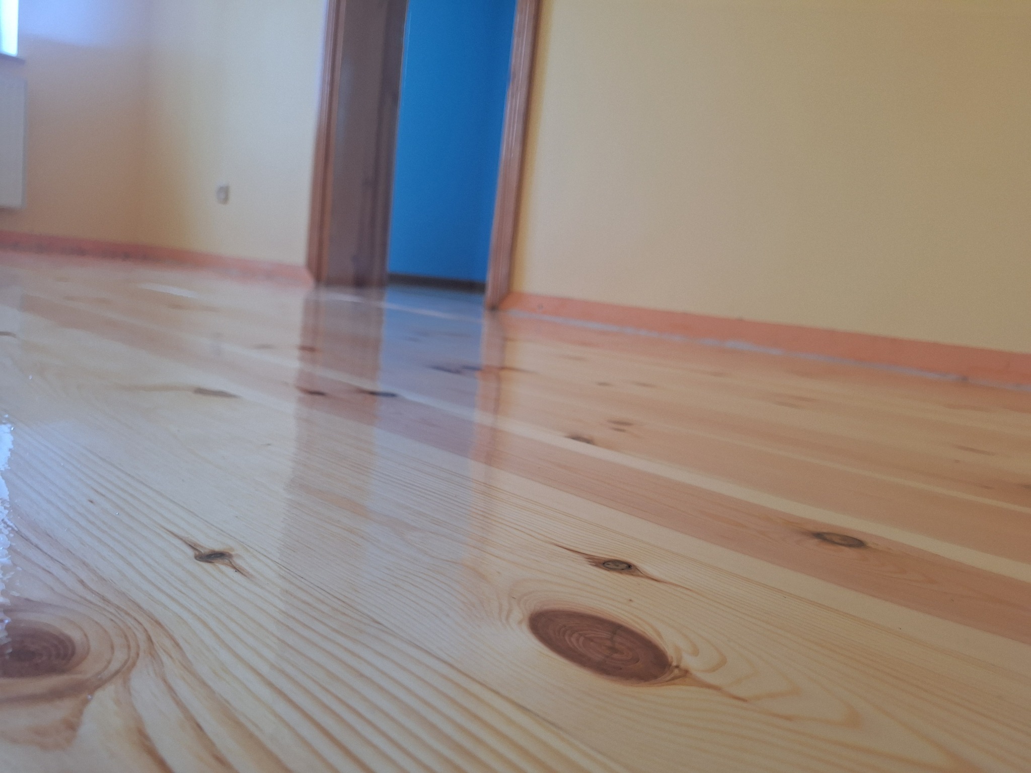 Błyszcząca, świeżo polakierowana podłoga z jasnego drewna z widocznymi słojami i sękami, odbijająca światło, z fragmentem drzwi w tle. Perspektywa z niskiego kąta.