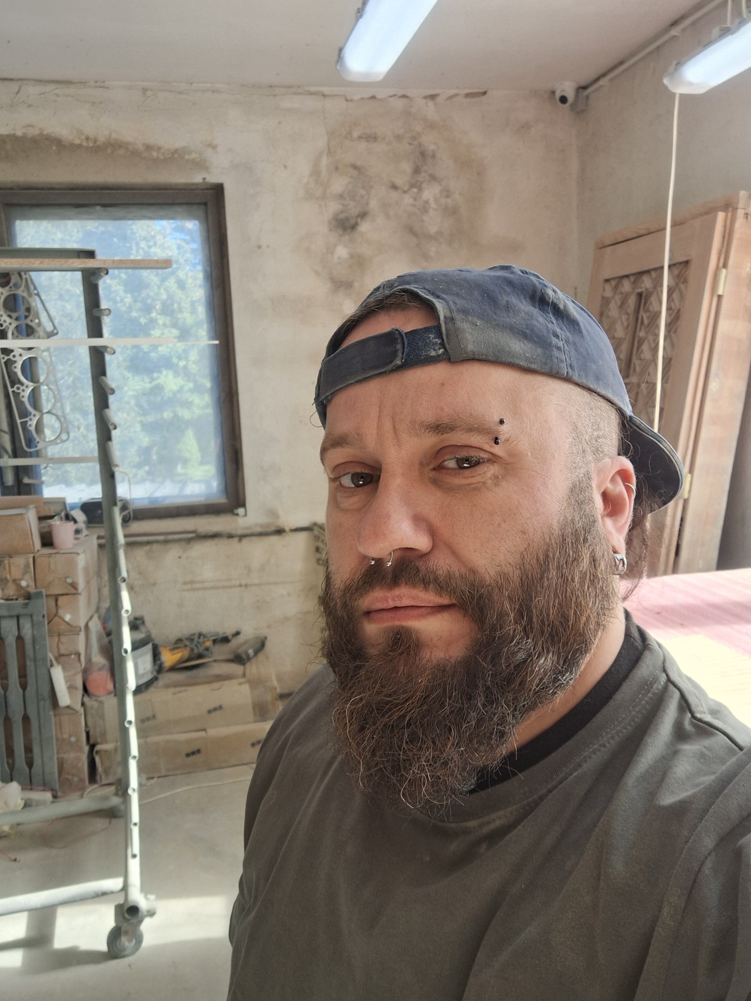 Portret brodatego mężczyzny w czapce na tle warsztatu z oknem, narzędziami i elementami drewnianymi. Widoczna kamera przemysłowa.