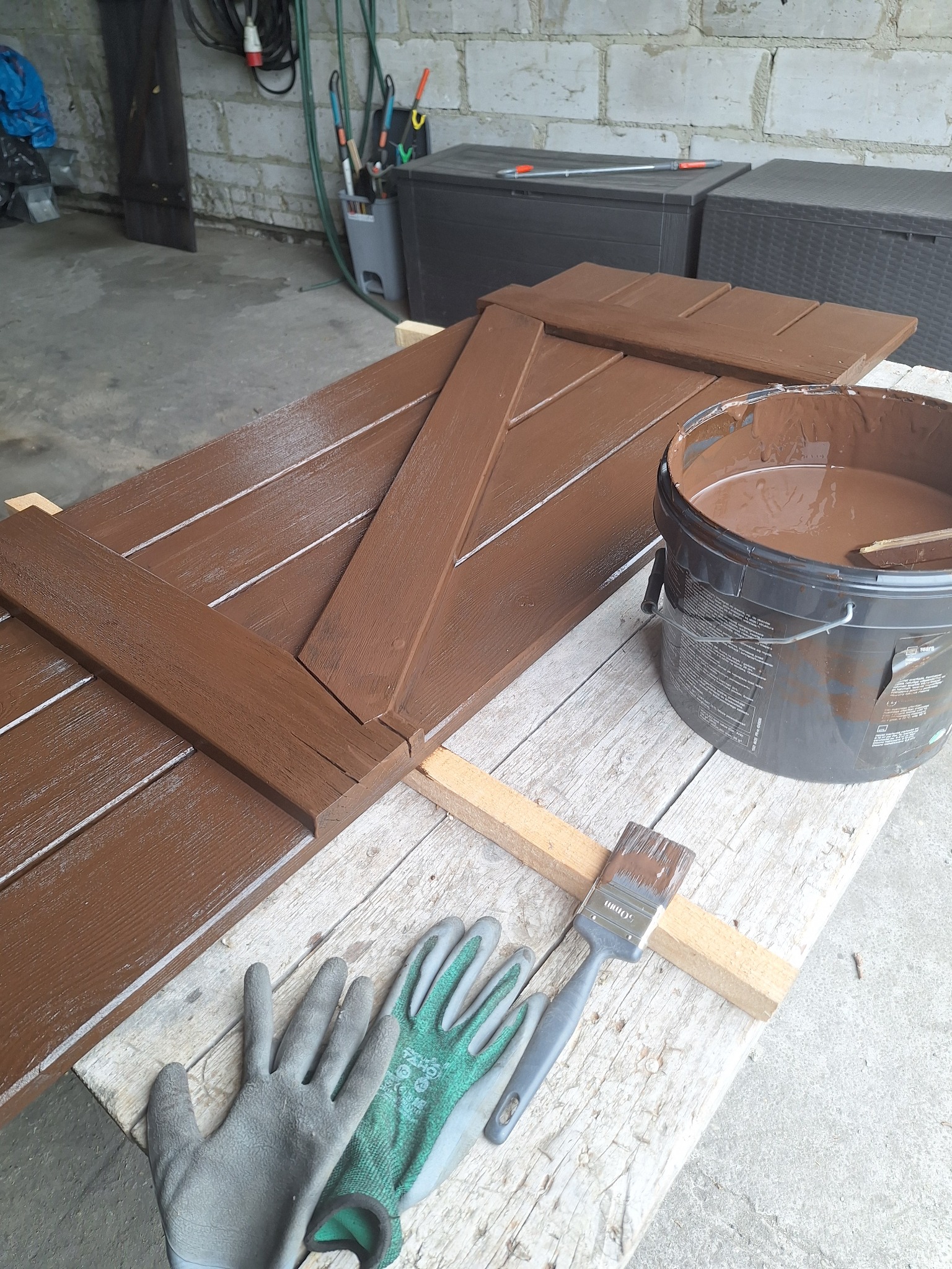 Pomazana brązową farbą drewniana konstrukcja drzwi na warsztatowym stole, obok puszka farby, pędzel i rękawice robocze. Widoczne ślady świeżego malowania.