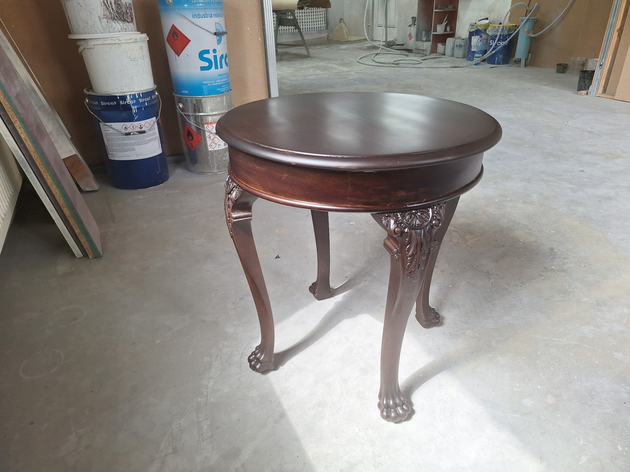 Okrągły, ciemnobrązowy stolik kawowy z rzeźbionymi nogami w warsztacie. W tle widoczne puszki z farbami i inne narzędzia stolarskie. Blat stolika odbija światło.