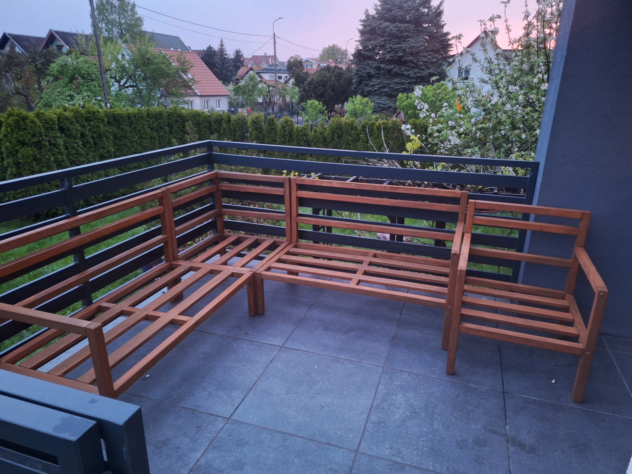 Drewniany, modułowy narożnik ogrodowy bez tapicerki na balkonie z szarymi płytkami. W tle zieleń i zabudowa jednorodzinna. Ujęcie z góry.