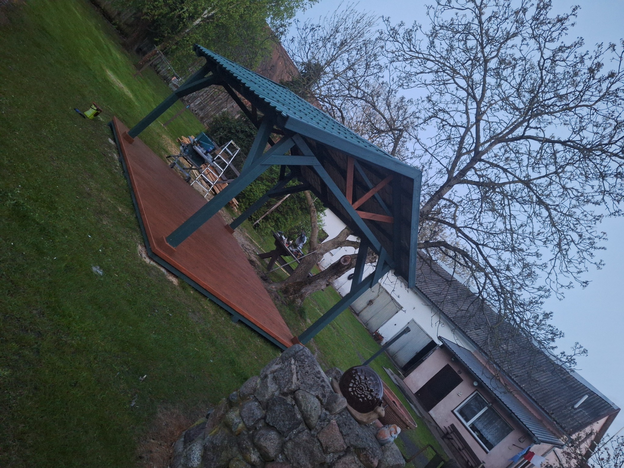 Drewniana altana z zielonym dachem na brązowym podeście, otoczona zielenią i kamiennym murkiem. Widoczne narzędzia i drabina, sugerujące świeży montaż.