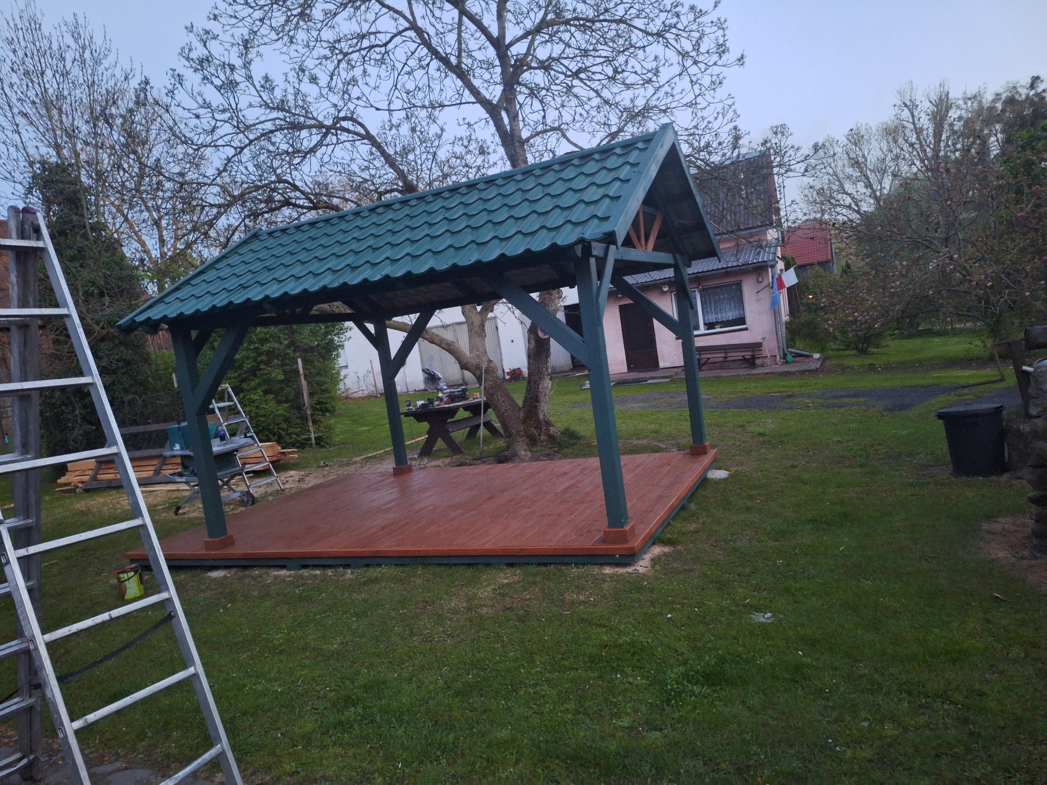 Zadaszona drewniana konstrukcja tarasowa z zielonym dachem na trawniku, widok z boku, drabina oparta o konstrukcję, w tle dom i drzewa.