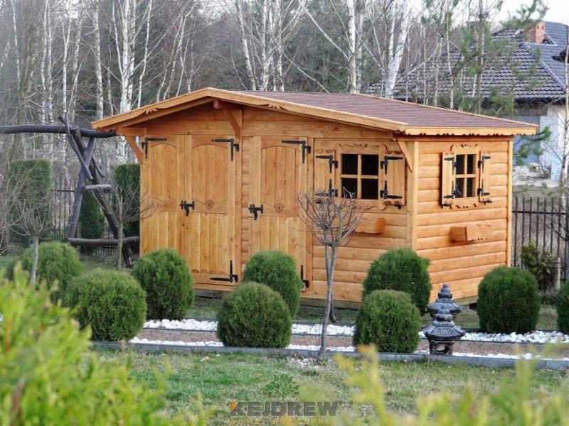 Drewniana altana ogrodowa z dwuskrzydłowymi drzwiami i oknami z okiennicami, otoczona kulistymi krzewami i białymi kamykami, z widocznym elementem huśtawki w tle.