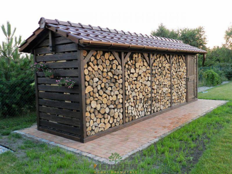 Drewniana wiata na drewno opałowe z brązowym dachem krytym dachówką, z małą furtką i doniczkami z kwiatami, ustawiona na ceglanym chodniku w ogrodzie.