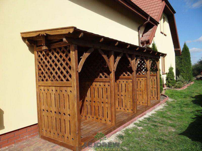 Drewniana altana na drewnianej podłodze, przylegająca do beżowej ściany domu z czerwonym dachem, otoczona zielonym trawnikiem i tujami.