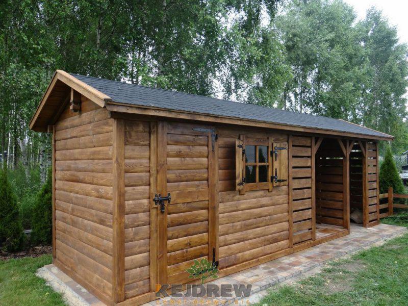 Drewniany domek narzędziowy z czarnym dachem, zamykanymi okiennicami i otwartą wnęką na drewno, umieszczony na wybrukowanym podłożu, widoczny na tle drzew.