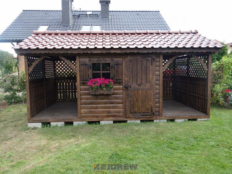 Drewniana altana ogrodowa z kratownicami, czerwonym dachem i skrzynką z różowymi kwiatami pod oknem, umieszczona na betonowych bloczkach, na tle domu z ciemną dachówką.