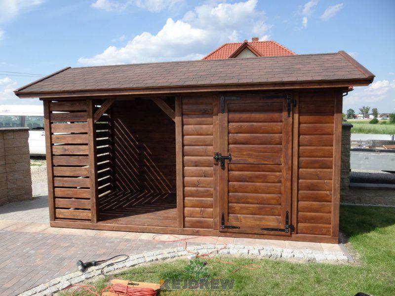 Drewniana drewutnia z zamykanymi drzwiami i otwartą przestrzenią na drewno, z brązowym dachem z gontu bitumicznego, stojąca na kostce brukowej z obrzeżem z kamienia naturalnego, widoczna w tle...