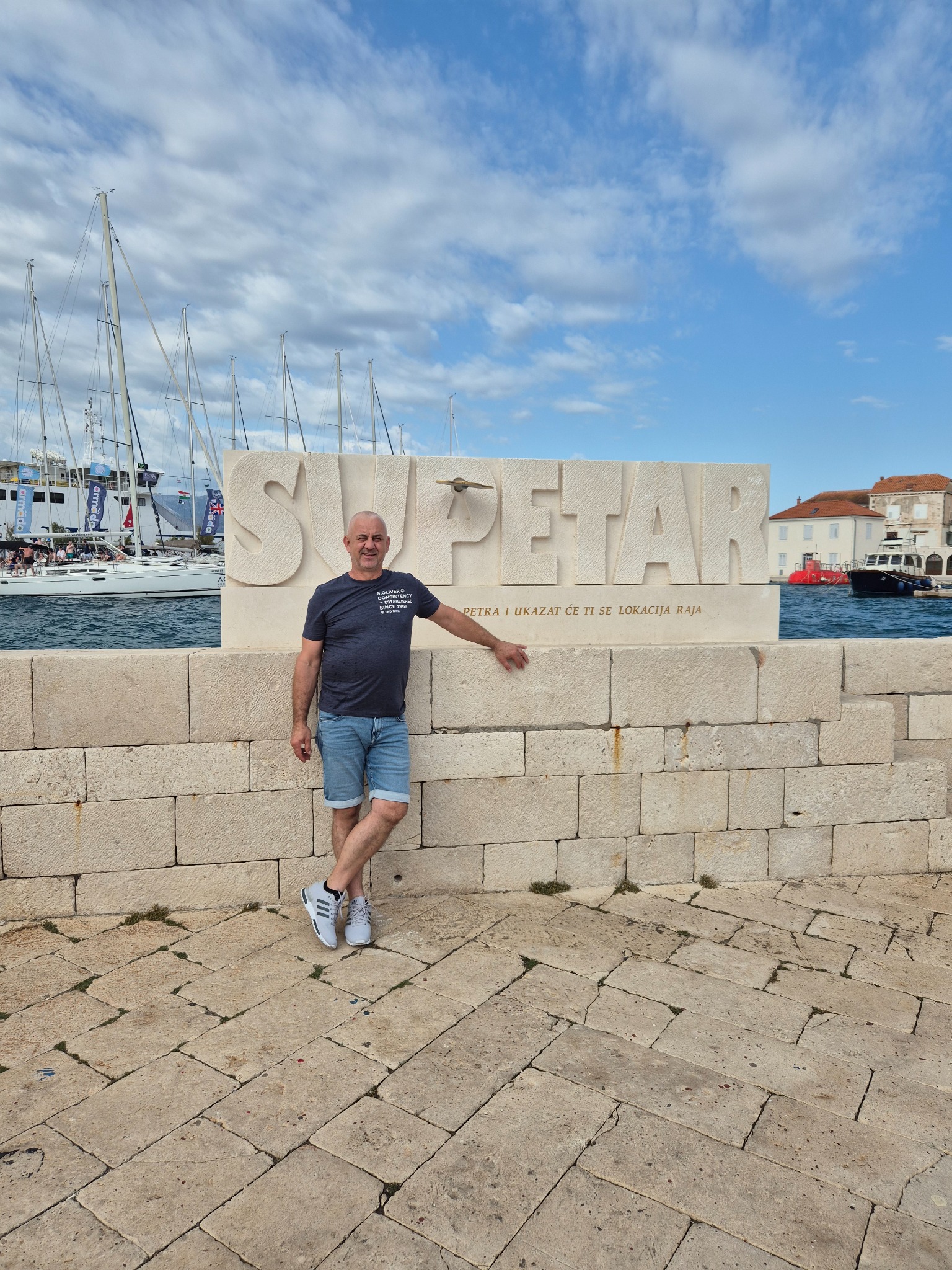 Mężczyzna opiera się o kamienny napis 'SUPETAR' na tle morza z żaglówkami. Jasne niebo z chmurami, kamienna nawierzchnia i budynki w oddali.