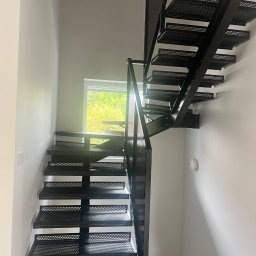 MS Metal Technik - Nowoczesne, ażurowe schody metalowe w jasnym wnętrzu. Minimalistyczny design z perforowanymi stopniami i prostą balustradą. Widok z dołu, okno w tle.