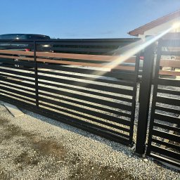 MS Metal Technik - Nowoczesna brama wjazdowa z czarnych, poziomych profili i drewnianymi akcentami, na tle domu i samochodu, widziana z bliska na żwirowym podłożu.