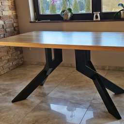 MS Metal Technik - Stół z blatem z jasnego drewna i czarnymi, metalowymi nogami o geometrycznym kształcie. W tle ceglana ściana i okno z roślinami doniczkowymi.