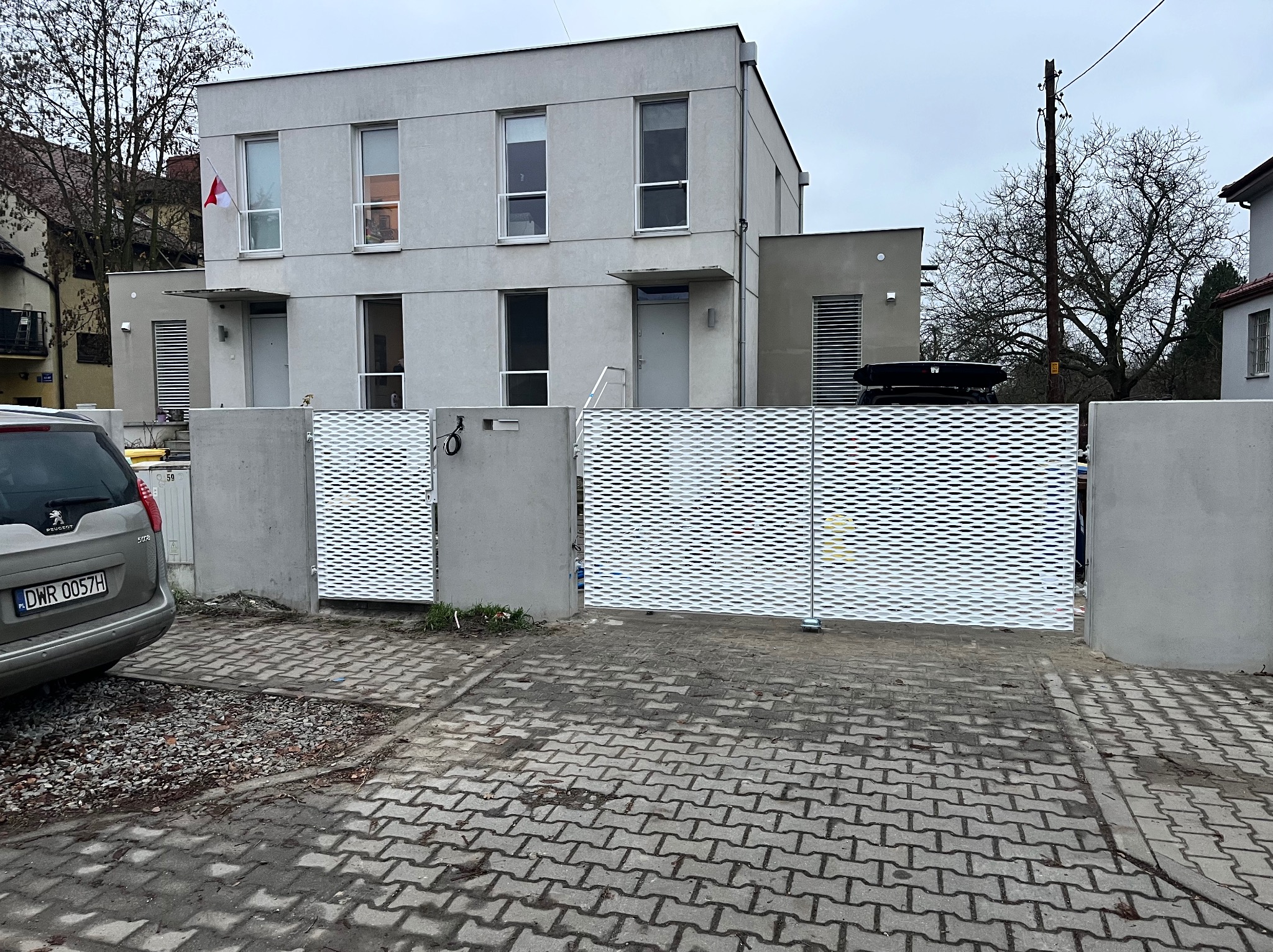 Nowoczesna, ażurowa brama wjazdowa z metalu, osadzona na betonowych słupkach, na tle dwukondygnacyjnego domu w minimalistycznym stylu. Widoczny fragment samochodu.