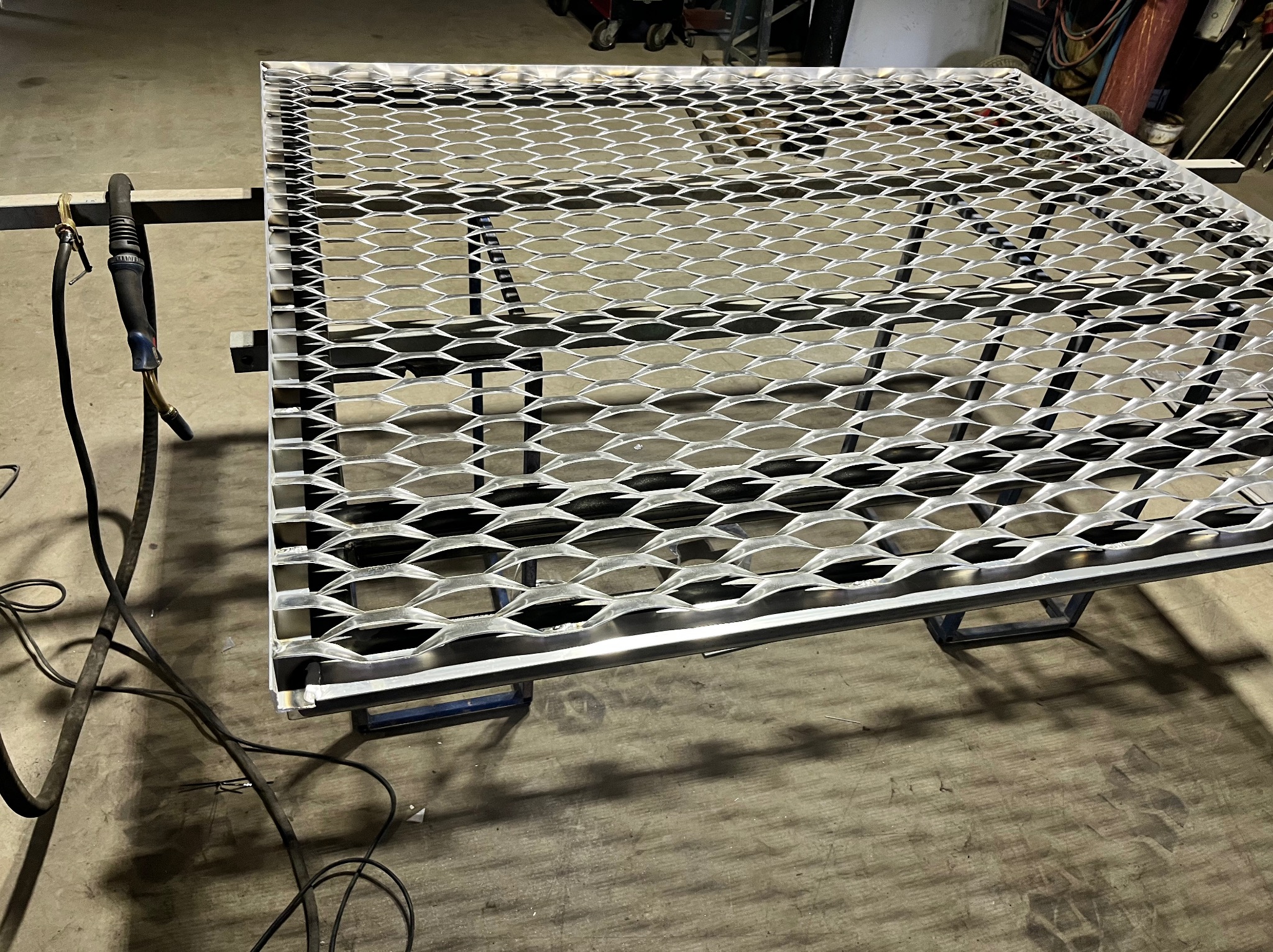 Metalowa krata z romboidalnymi oczkami, spawana na ramie, leżąca na warsztatowym stole. Widoczny przewód spawalniczy i surowa posadzka warsztatu.