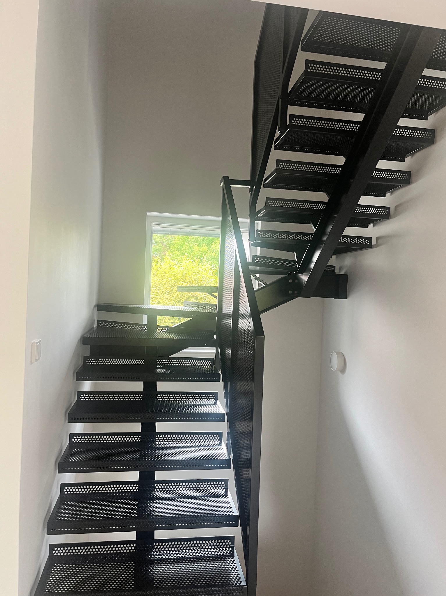 Nowoczesne, ażurowe schody metalowe w jasnym wnętrzu. Minimalistyczny design z perforowanymi stopniami i prostą balustradą. Widok z dołu, okno w tle.