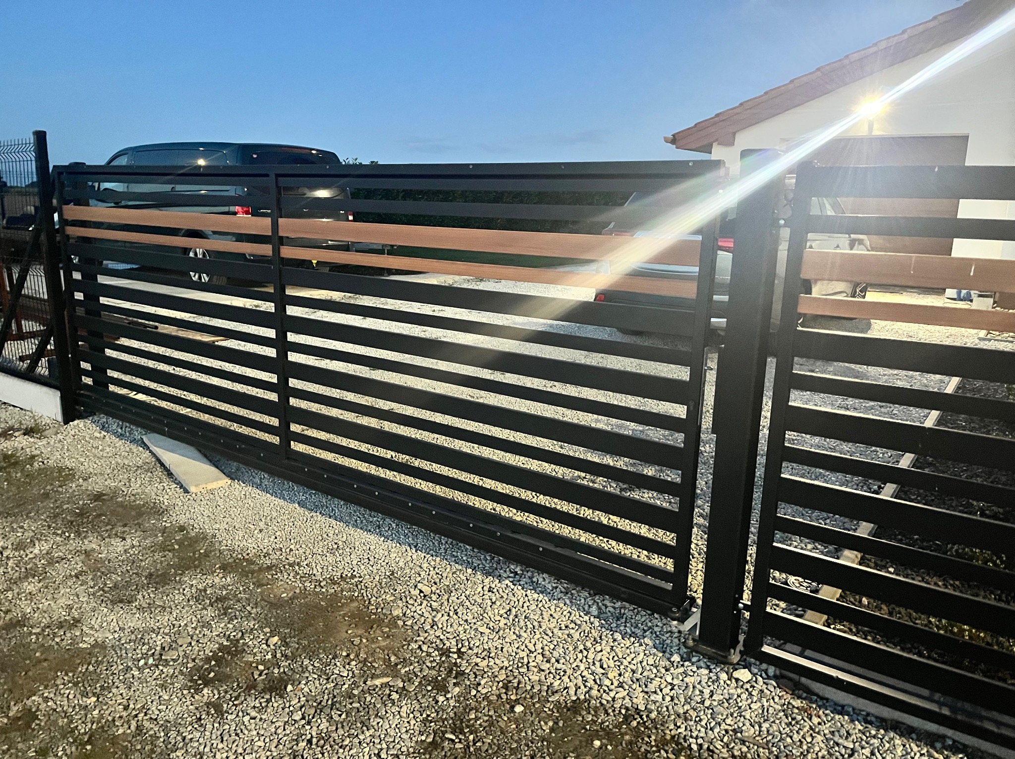 Nowoczesna brama wjazdowa z czarnych, poziomych profili i drewnianymi akcentami, na tle domu i samochodu, widziana z bliska na żwirowym podłożu.