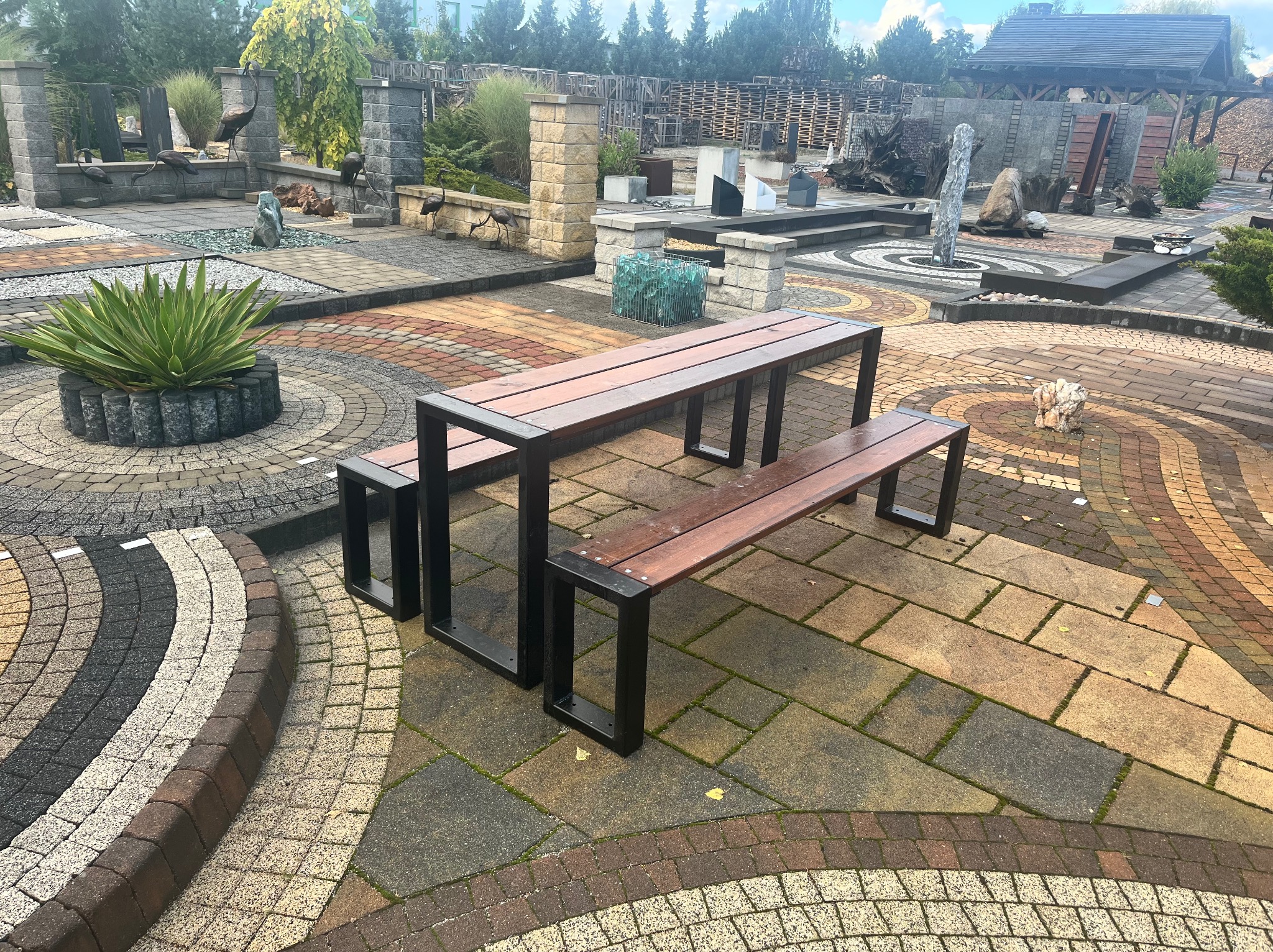 Zestaw mebli ogrodowych: stół z dwiema ławkami na tle ekspozycji kostki brukowej w różnych kolorach i wzorach. Widoczne elementy małej architektury ogrodowej.