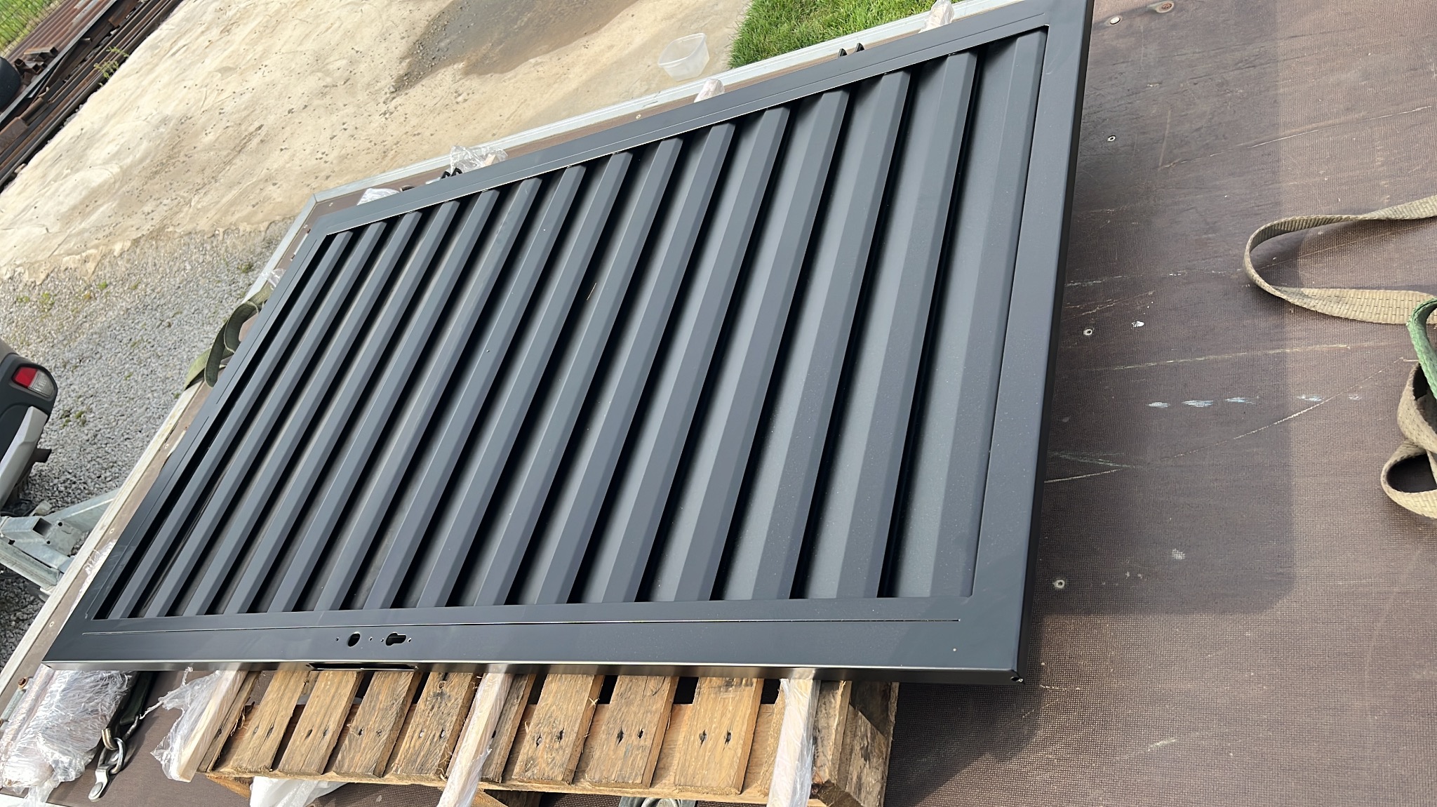 Ciemnoszara, metalowa brama wjazdowa z pionowymi panelami, leżąca na drewnianej palecie. Widoczne otwory montażowe i fragment trawy w tle. Ujęcie z góry.