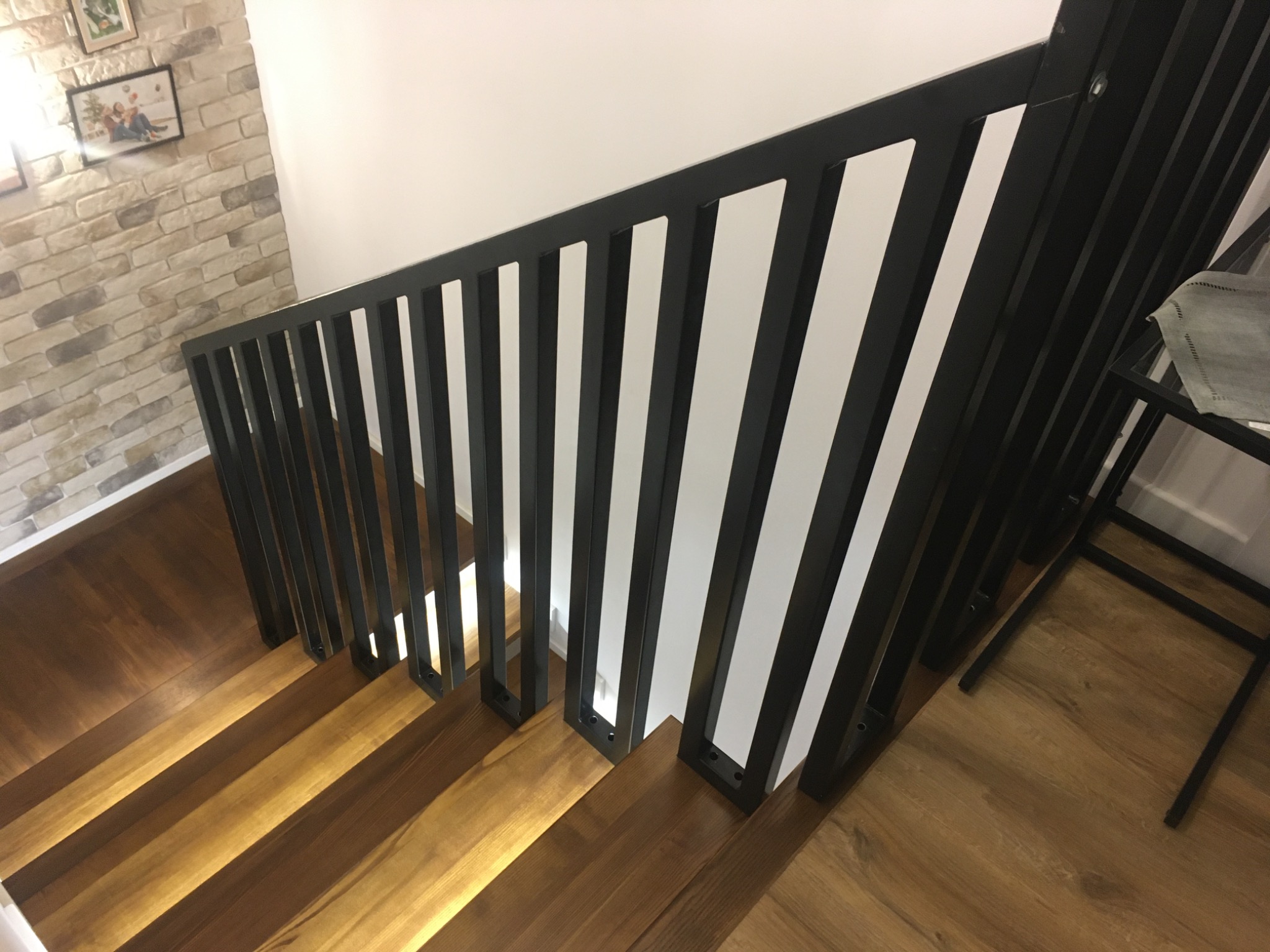 Nowoczesne schody z drewnianymi stopniami i czarną, metalową balustradą z pionowymi szczebelkami. Widok z góry na dół, oświetlenie podkreśla strukturę drewna.